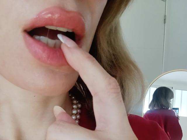 Los labios carnosos y rojos de MissFrenchy están ligeramente entreabiertos, con su dedo, cuidadosamente arreglado, tocando el labio inferior. Tiene la piel pálida, el cabello rubio y un collar de perlas. Se puede ver su reflejo..