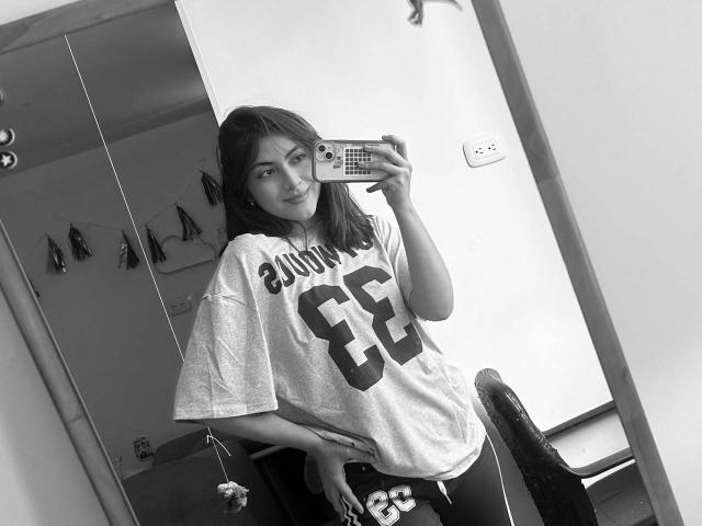 SpencerLe, con un busto de tamaño medio, vistiendo una camiseta deportiva holgada con el número 65, tomando una selfie frente al espejo en una foto en blanco y negro, sosteniendo un teléfono con una funda a cuadros..