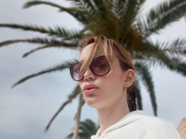 MissFrenchy con gafas, labios rosados, piel clara, cabello rubio, sudadera blanca con capucha, pequeños aretes dorados, palmera en el fondo, cielo nublado, mirando ligeramente hacia abajo, expresión suave..