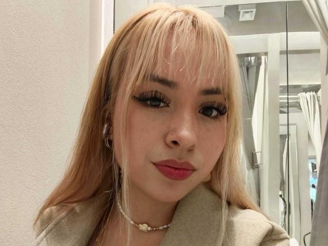BerlyQueen has blonde hair, long eyelashes, pink lipstick, and a cute smile. She's wearing a beige coat and a pearl necklace. Her nose is pierced.