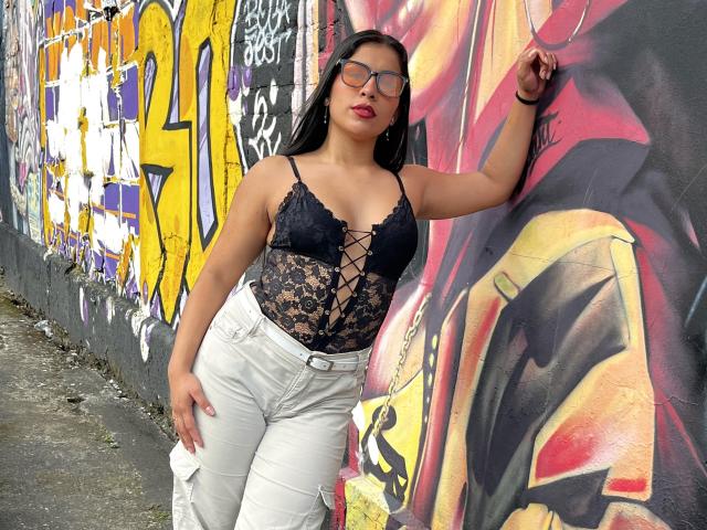 MelodyRoss with black lace bodysuit, white jeans, and glasses, leaning against graffiti wall, her hand on the wall, her ass slightly pressed against it.
