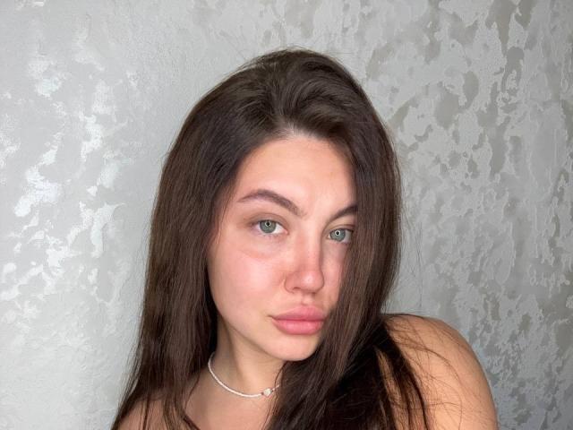 Ines Gueningsman tiene el pelo largo y castaño, ojos verdes, labios rosados y un collar discreto. Sus hombros desnudos son visibles. Está mirando directamente a la cámara con un ligero puchero..