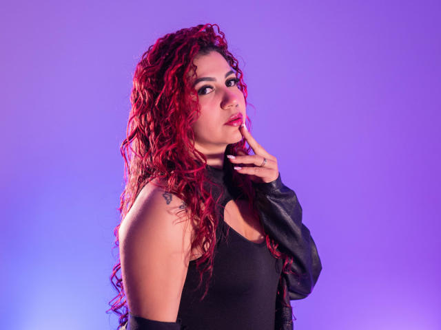 Antonela Riveiro, con capelli rossi ricci, top nero senza maniche e giacca di pelle nera, che tocca le labbra con un dito, sfondo viola, tatuaggio sulla spalla sinistra..