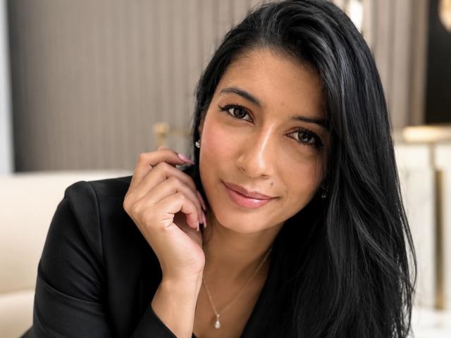 JasminKooper ha lunghi capelli neri, occhi marroni e un leggero sorriso. Indossa una maglietta nera, tocca la guancia con la mano destra e ha una collana..