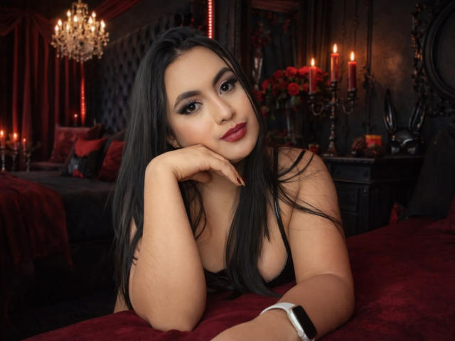 Scarlett Rogers, con lunghi capelli neri, rossetto rosso e trucco occhi scuro, appoggia la testa sulla mano, mostrando il seno, in una camera da letto scura, illuminata di rosso e con delle candele..
