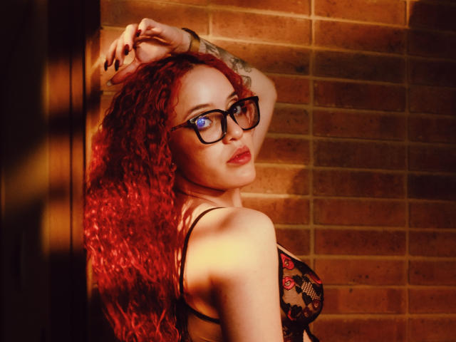 LilJane, con cabello rizado rojo, gafas y un sujetador de encaje negro, mirando por encima del hombro, con una mano en la pared, un tatuaje en el brazo, fondo de ladrillo naranja, sexy, segura de sí misma, coqueta..