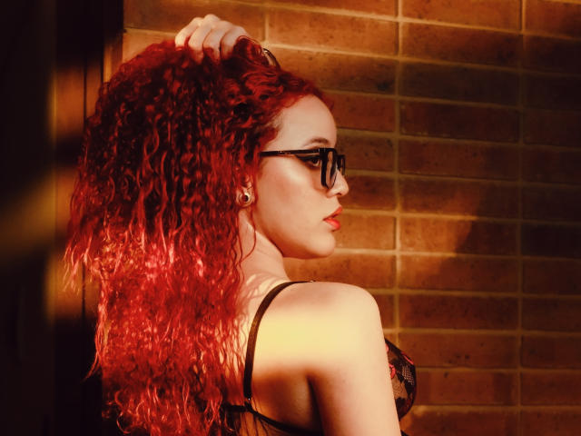 LilJane con capelli rossi ricci, occhiali neri e reggiseno di pizzo nero, mano tra i capelli, profilo laterale, luce solare, muro di mattoni, piercing alle orecchie, sexy, pelle chiara, labbra rosse..
