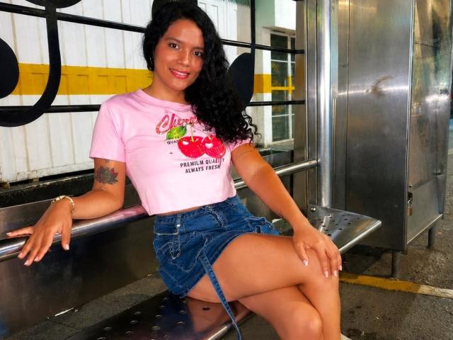 Amily Harlow, con cabello negro rizado, camiseta rosa con la palabra "Cherry", y falda vaquera, sentada en un banco de metal, mostrando un tatuaje en el brazo derecho, sonriendo, con las piernas cruzadas, atractiva, linda, de piel morena..