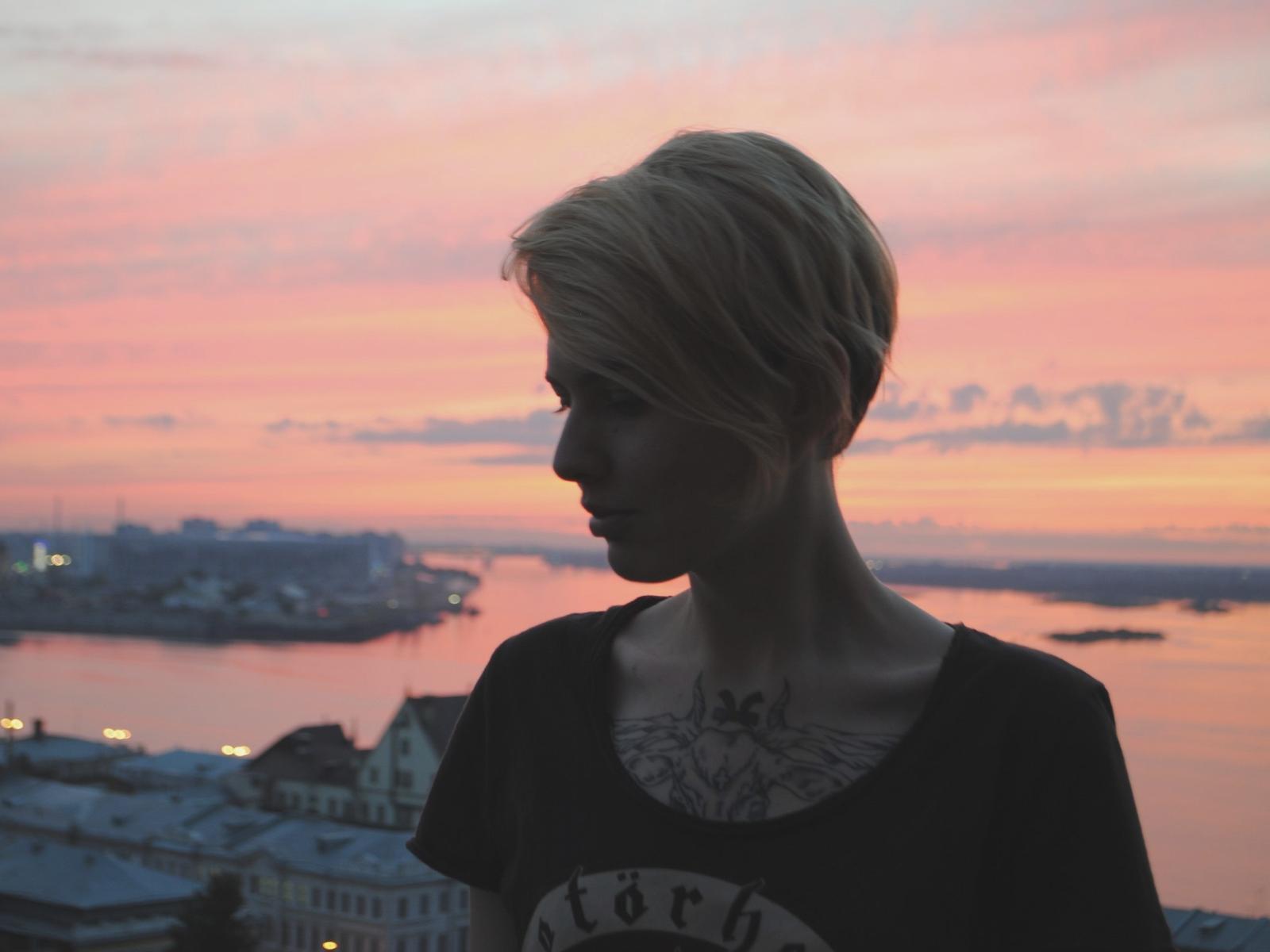 Stacey69 hat kurze, blonde Haare, ein schwarzes T-Shirt und ein Brust-Tattoo und steht im Schatten vor einem rosa und orangefarbenen Sonnenuntergang. Ihre kleinen Brüste sind sichtbar..