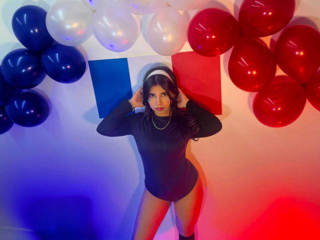 CelesteMorel with dark hair, wearing a black long-sleeve leotard, standing against red, white, and blue balloons, hands in her hair, looking at the camera, legs apart.