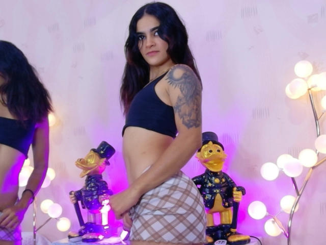 Une personne, cheveux foncés, bras tatoué, soutien-gorge de sport noir et pantalon à carreaux, debout à côté de deux soldats jouets, avec des lumières violettes et blanches en arrière-plan..