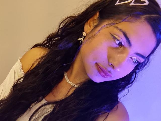 AngelAlba with wavy black hair, wearing a white off-shoulder top, glittery eyeliner, diamond earrings, and a diamond necklace, has purple-tinged skin and a small, shiny lip gloss.