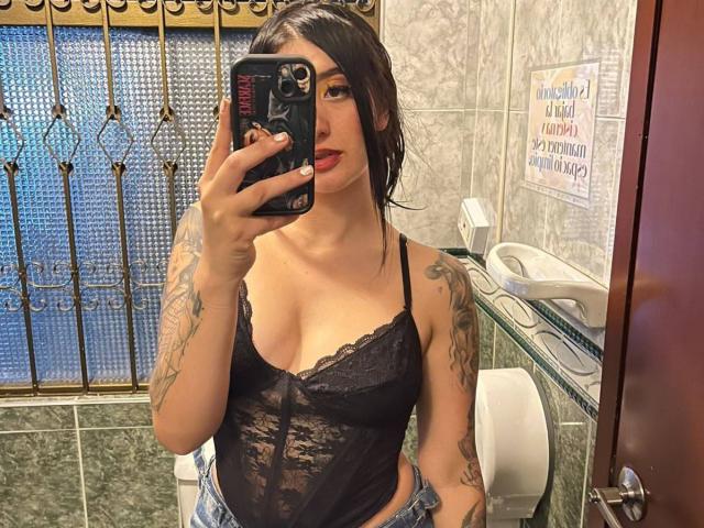 TifanyDiaz met tatoeages, een zwarte kanten top, rode lipstick en een spijkerbroek, maakt een spiegelzelfportret in een badkamer, waarbij haar kleine borsten zichtbaar zijn en ze een zwarte telefoon vasthoudt..
