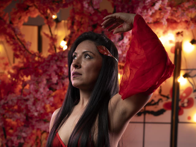 EllaRousse mit langem, schwarzem Haar, einem roten Kleid und passenden Armstulpen, die ihren rechten Arm hebt und dabei ihre Brüste zur Schau stellt, vor einem roten und rosa Blumenhintergrund, mit nach oben gerichtetem Blick..