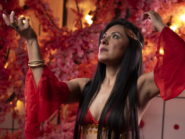 EllaRousse, com longos cabelos pretos, mangas vermelhas transparentes, pulseira dourada e sutiã vermelho, com os braços levantados, olhando para cima, em um cenário floral vermelho e rosa. Seus seios estão visíveis..