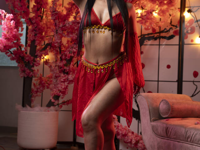 EllaRousse vestindo lingerie vermelha com um cinto de corrente dourada, mostrando os seios e uma perna, em pé com os braços levantados, cabelo preto, fundo com flores de cerejeira rosa e um sofá rosa..