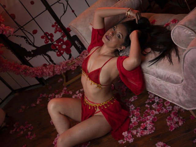 EllaRousse, con cabello oscuro, traje de baño rojo y una blusa roja transparente, sentada en el suelo de madera, rodeada de pétalos rosas, con los brazos por encima de la cabeza, las piernas separadas, mirando a la cámara, sexy..