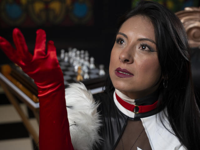 EllaRousse with dark hair, red gloves, black leather outfit, white fur, red collar, looking up, hand raised, purple lipstick, brown eyes, in a photo with a chessboard.