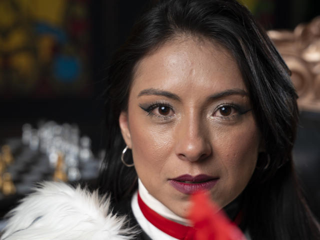 EllaRousse, mit dunklem Haar und großen Ohrringen, hat braune Augen, dezentes Make-up und rosafarbenen Lippenstift. Sie trägt einen weißen Pelzkragen und ein rotes Band. Ihr Gesicht ist scharf..
