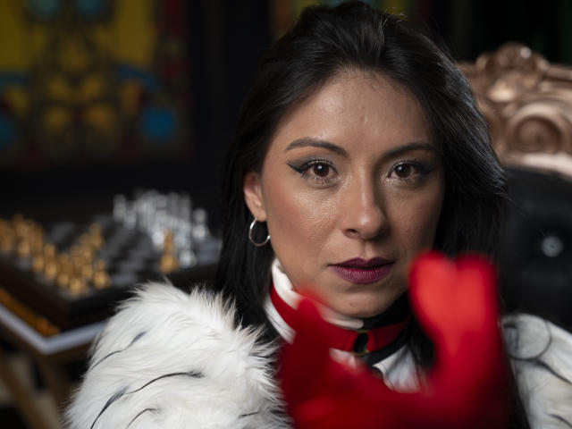 EllaRousse, con cabello oscuro, ojos marrones y lápiz labial rojo, lleva un collar de piel blanca y un collar de cuentas rojas, tiene una expresión seria y usa aros grandes, con un guante rojo en primer plano..
