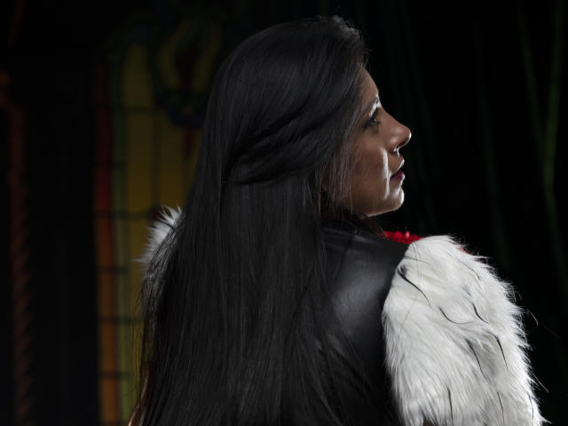 EllaRousse, avec ses longs cheveux noirs, son gilet en cuir noir et ses manches en fourrure blanche, montrant son profil avec un ruban rouge sur sa poitrine. Fond noir et velouté..