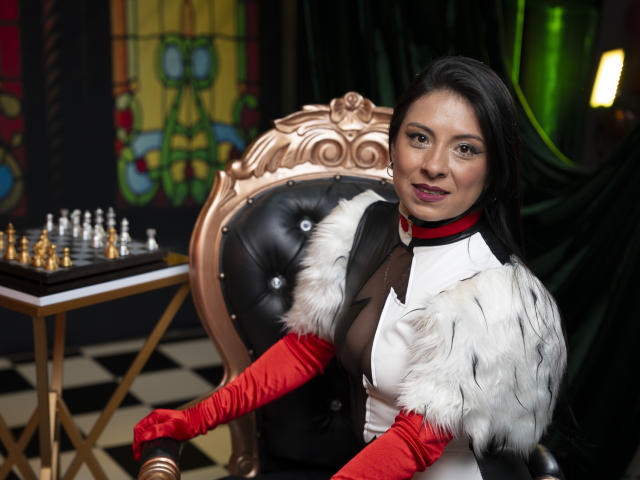 EllaRousse, with dark hair and red gloves, sits in an ornate chair, wearing a white dress with black and white fur, showing her tits, near a chessboard.