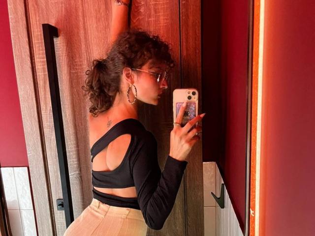 Alyce Erway, com cabelos castanhos cacheados, óculos e brincos de argola, vestindo uma blusa preta com recortes e calças bege de cintura alta, tirando uma selfie no espelho com o braço direito levantado..