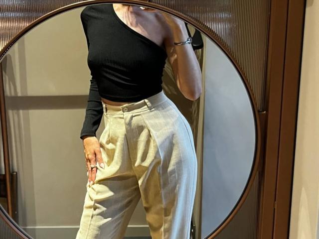 AlyceErway con una blusa negra de un solo hombro, mostrando su pequeño trasero en pantalones holgados de color gris claro y de cintura alta, de pie frente a un espejo redondo..