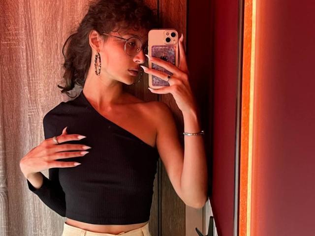AlyceErway, con i capelli ricci, gli occhiali, una maglietta nera con una spalla scoperta e degli orecchini, sta scattando un selfie allo specchio, mostrando la mano sul petto, con un telefono nell'altra mano..