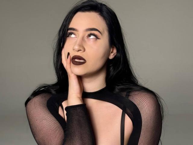 Mevrouw Mary Lori, met donkere lipstick, een zwarte kanten top en lang zwart haar, kijkt omhoog met haar hand op haar wang, waarbij haar borsten zichtbaar zijn door de uitsparingen in het kant, en ze ziet er sexy en verleidelijk uit..