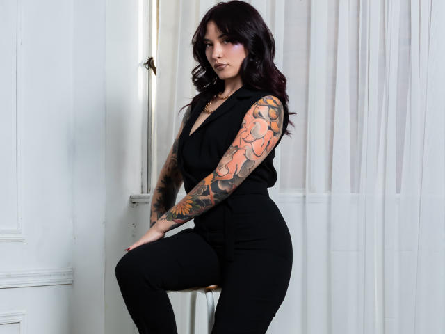 AkiraNishimura69, getatoeerde armen, zwart mouwloos topje, zwarte broek met hoge taille, zittend, witte muur, doorschijnend gordijn, donker golvend haar, paars oogschaduw, serieuze uitdrukking, sexy, tatoeages zichtbaar op de rechterarm..