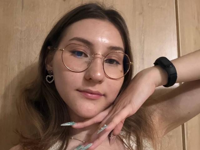 Lenore Schontz tem uma pele luminosa, usa óculos redondos, uma pulseira preta e unhas longas e brilhantes. Ela está tocando o queixo com a mão direita, mostrando um sorriso discreto..