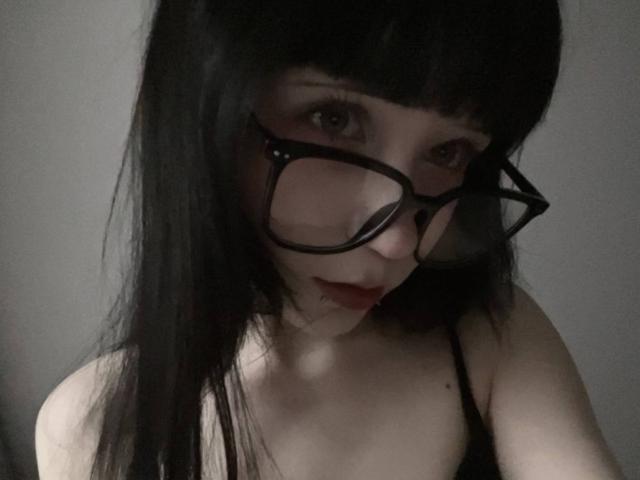 Nancy Turpin, com óculos pretos, pele clara e cabelo preto liso, olhando para baixo com batom vermelho, vestindo uma blusa preta de alças finas, mostrando seus pequenos seios..