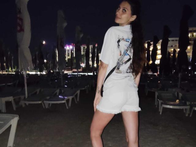 DanaWaw, con lunghi capelli scuri, una t-shirt bianca con grafica e pantaloncini bianchi sfilacciati, è in piedi con la schiena rivolta alla telecamera, guardando oltre la spalla, mostrando il sedere e un tatuaggio sul braccio sinistro..