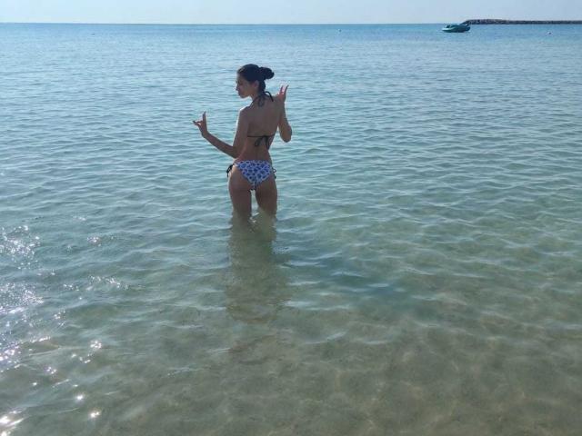 DanaWaw steht im klaren Wasser, mit dem Rücken, und trägt einen blau-weißen Bikini mit Muster, wobei ihr Hintern zu sehen ist. Ihre Hände sind erhoben, und sie schaut über ihre Schulter zurück..