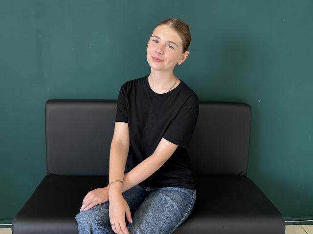 AdelinaStar, con piccoli seni, figura snella e pelle chiara, indossa una maglietta nera e jeans blu, seduta su una panchina nera, sorride dolcemente, capelli castani raccolti in una coda di cavallo..