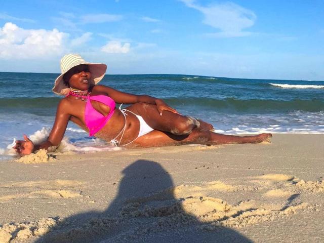 KuteFatPussy is a dark-skinned woman with big boobs, wearing a pink bikini top, white bottom, and sunhat, lounging on a sandy beach with her ass and toes in water.