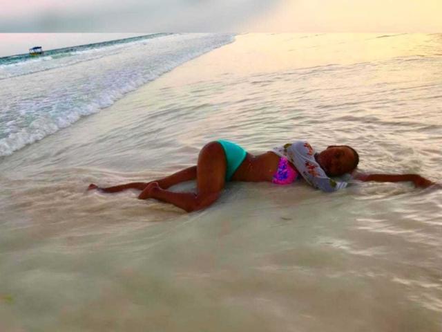 KuteFatPussy, à la peau foncée, allongée dans une eau peu profonde, portant un bas de maillot de bain turquoise et un haut rose, violet et bleu, avec les bras tendus et les jambes légèrement écartées..