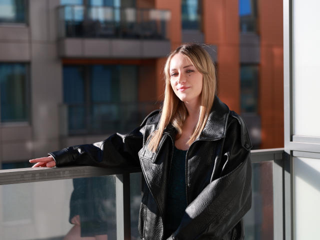 EvelynBlond, con lunghi capelli biondi, giacca di pelle nera e top scuro, appoggiata a un balcone di vetro, con la luce del sole sul viso, espressione calma, sullo sfondo un ambiente urbano..