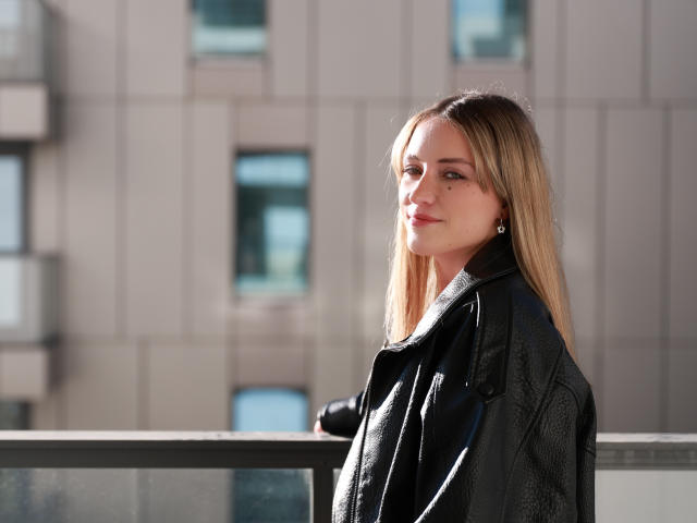 EvelynBlond, avec ses cheveux blonds, portant une veste en cuir noir, se tient près d'un balcon en verre, regardant sur le côté avec un léger sourire, la lumière du soleil sur son visage..