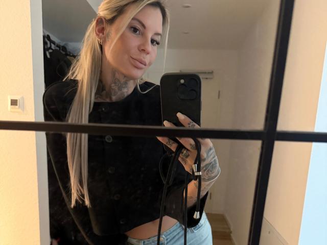 Siera, une jeune femme aux longs cheveux blonds, avec des tatouages, portant un crop top noir, tenant un téléphone, prenant un selfie devant un miroir, montrant ses seins et ses tatouages sur les bras, vêtue d'un jean bleu, avec un léger sourire, dans un intérieur..