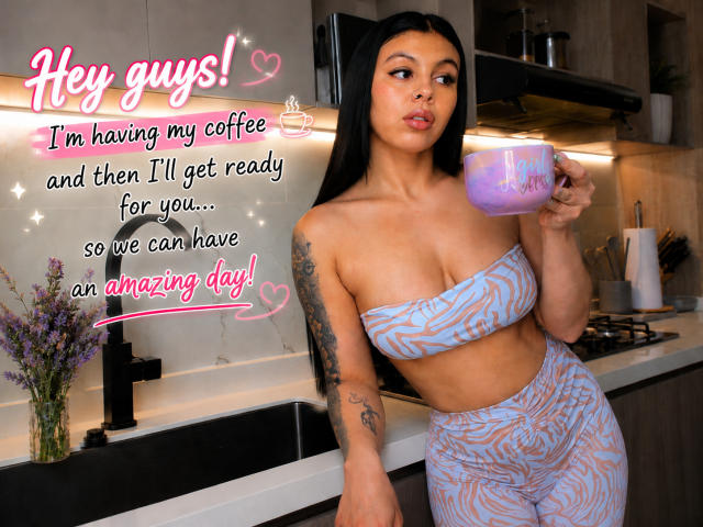 NenaBlack with long black hair, tan skin, and tattoos, holding a purple coffee mug, wearing a blue patterned tube top and matching high-waisted pants, standing in a kitchen.