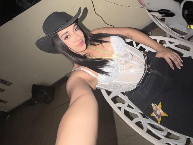 EmmaWaltter con un cappello nero, una camicetta di pizzo bianco e jeans neri, che mostra un selfie con un braccio esteso, rivelando il seno e una stella gialla sui pantaloni..