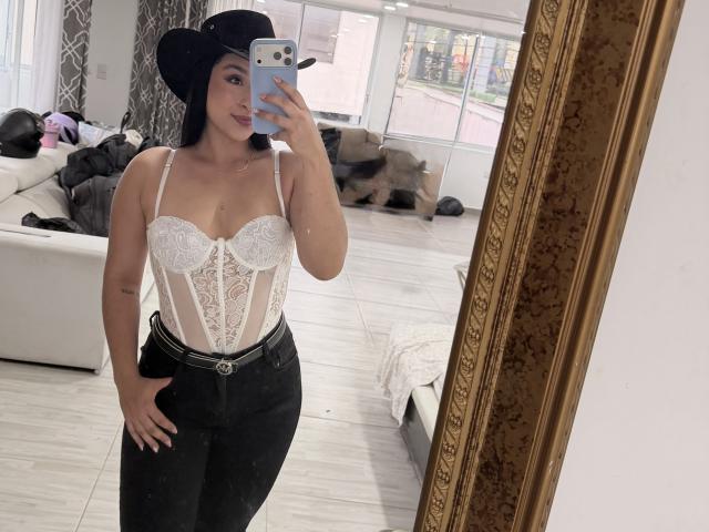 Emma Waltter trägt ein weißes Spitzen-Korsett, schwarze Hosen, einen schwarzen Cowboyhut, hält ein Telefon, macht ein Selfie im Spiegel, zeigt ihre Brüste, hat eine Hand auf ihrem Hintern, in einem modernen Raum..
