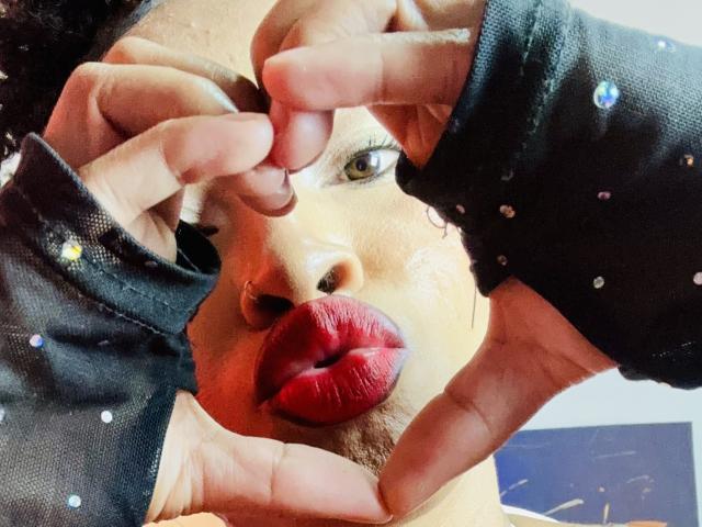 El rostro de Plaisir69, con su piel morena, labios rojos y guantes negros sin dedos adornados con brillos, enmarca sus ojos y nariz con los dedos. Tiene el cartílago de la nariz perforado y está mirando a la cámara..