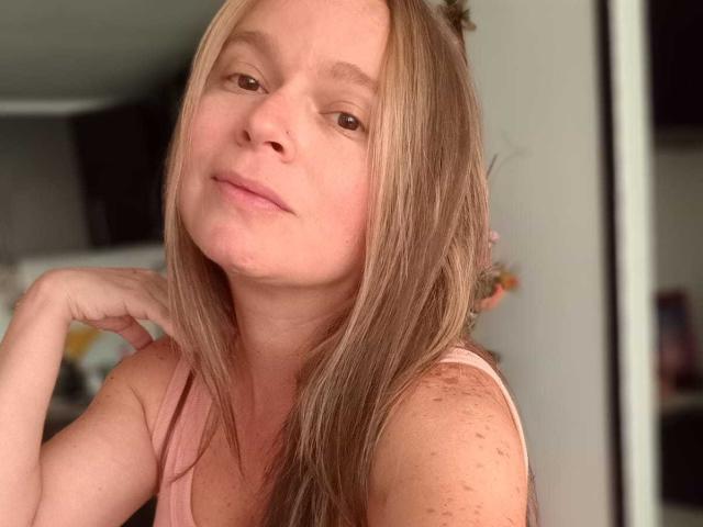 PamelaColin with blonde hair, pink tank top, freckled shoulders, relaxed expression, slightly tilted head, hand under chin, soft smile, casual bedroom setting.