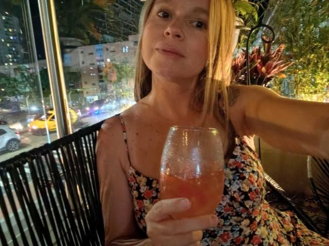 PamelaColin with blonde hair, floral dress, holding a pink cocktail, sitting in a dimly lit cafe at night, visible city lights and cars in the background.