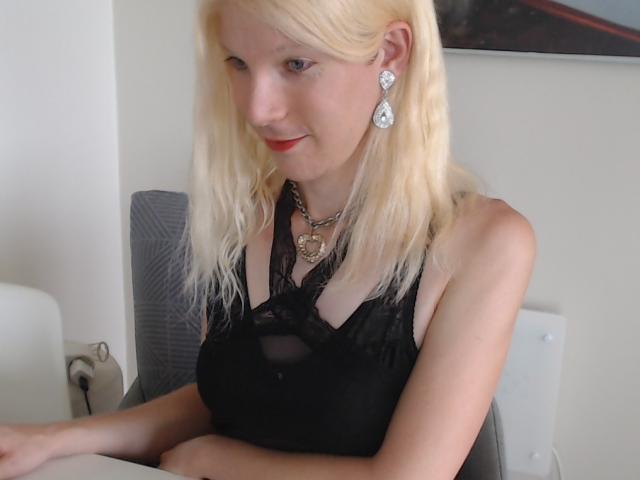 Natacha Bradley mit blonden Haaren, rotem Lippenstift, einem schwarzen Spitzenoberteil, das ihre kleinen Brüste zeigt, trägt eine Herz-Kette und Diamant-Ohrringe, sitzt auf einem Stuhl und lächelt..