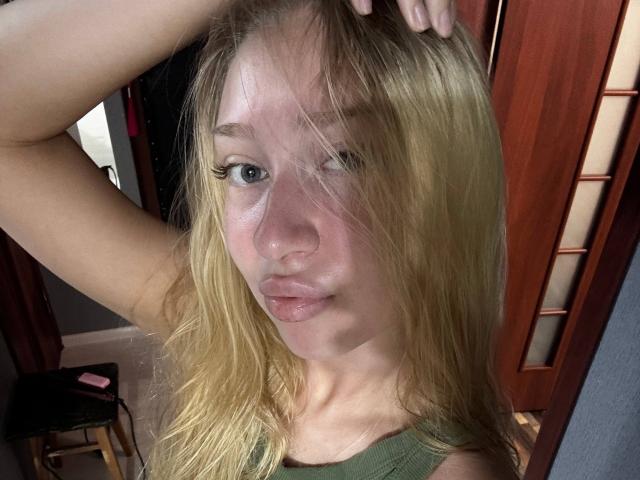 Joette Shigeta met blond haar, volle lippen, blauwe ogen, en één oog gedeeltelijk gesloten, met haar in haar handen, gekleed in een groene top, in een selfie met een houten deur op de achtergrond..