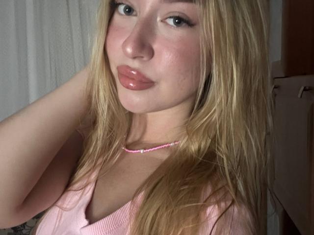 Joette Shigeta mit blonden Haaren, pinkem Lippenstift und einer pinken Halskette, trägt ein pinkes Oberteil, das ihre kleinen Brüste zeigt, und hat einen süßen, schmollenden Gesichtsausdruck auf einem Selfie..